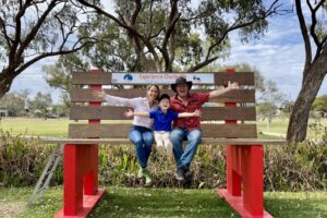 IMG_5041 A family sitting on a giant bench at Charleville in Outback Queensland, smiling with arms wide open, surrounded by gum trees and open parkland.