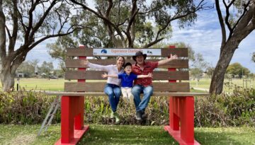 A family sitting on a giant bench at Charleville in Outback Queensland, smiling with arms wide open, surrounded by gum trees and open parkland.