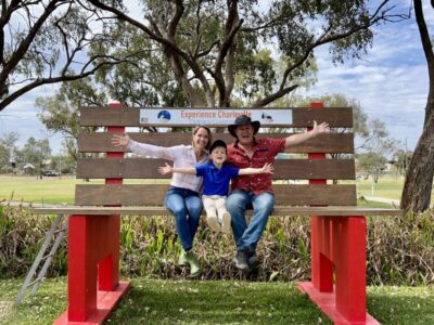 IMG_5041 A family sitting on a giant bench at Charleville in Outback Queensland, smiling with arms wide open, surrounded by gum trees and open parkland.