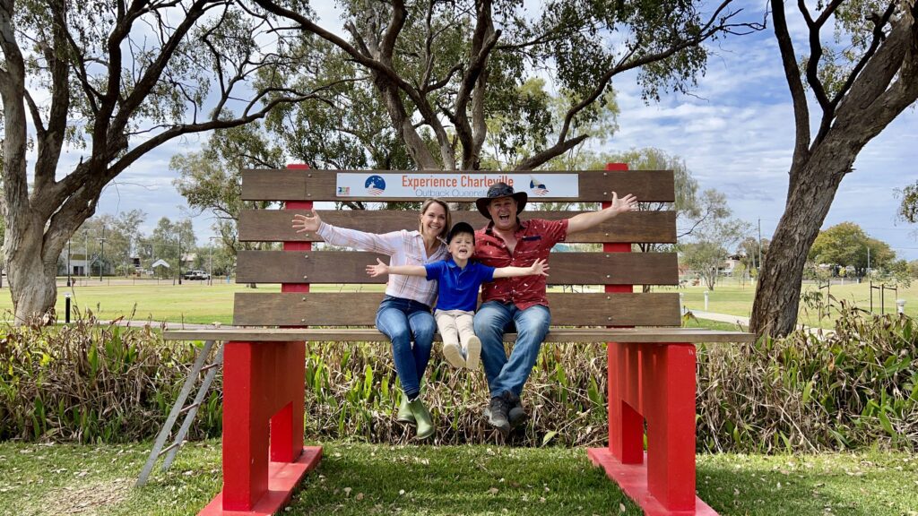 A family sitting on a giant bench at Charleville in Outback Queensland, smiling with arms wide open, surrounded by gum trees and open parkland.